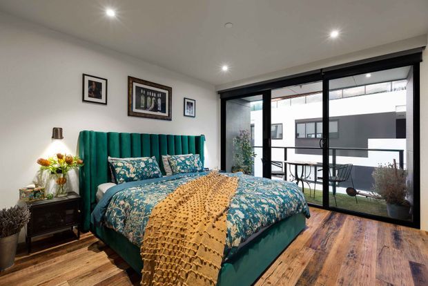 A stylish bedroom with a teal upholstered bed and matching teal bedding, layered with floral-patterned pillows and a mustard knitted throw. The room has warm timber flooring, white walls, recessed ceiling lights, framed artwork, a bedside table with lamp and flowers, and large black-framed glazed doors opening to a small balcony or courtyard area.