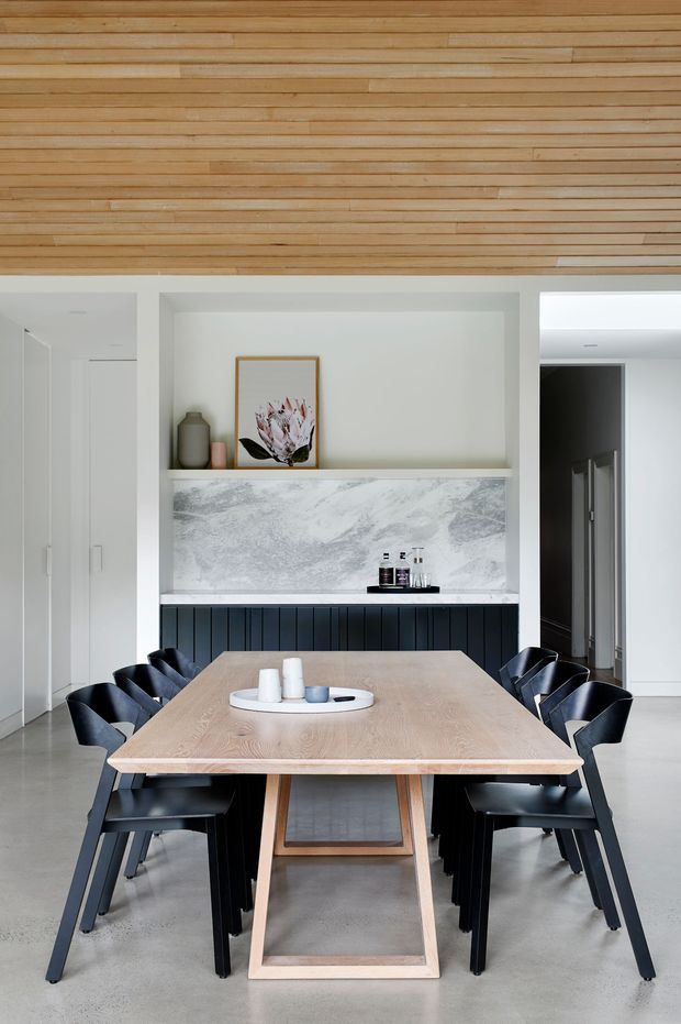 A contemporary dining room with a long light timber dining table surrounded by black dining chairs. The room features white built-in cabinetry, a recessed open shelf with understated decor, a grey-and-white stone-look feature splashback, dark vertical-panel joinery below, polished concrete flooring, and a striking timber slatted ceiling.