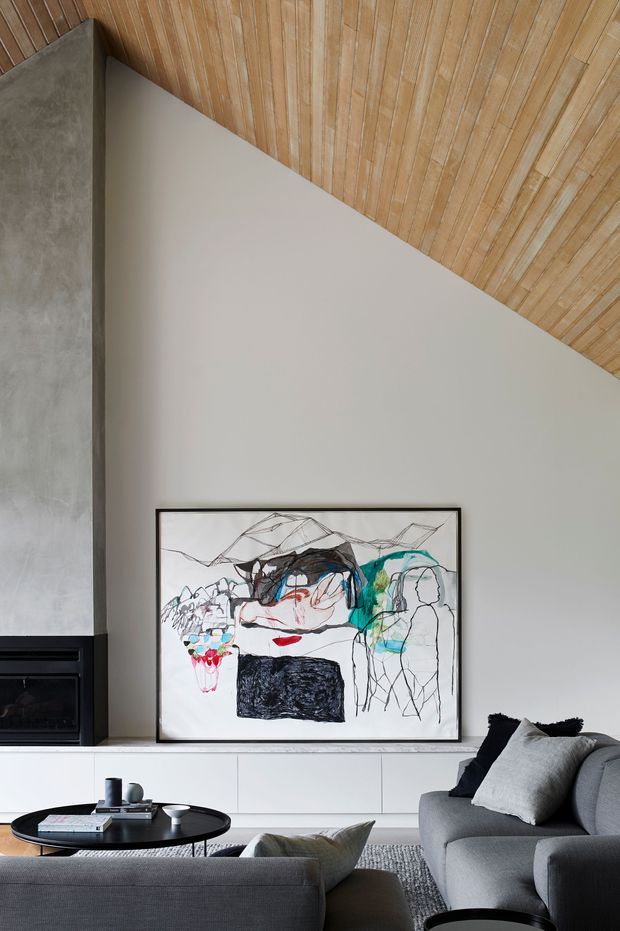 A contemporary living room with a tall sloping timber-lined ceiling, smooth pale walls, a dark concrete or stone fireplace surround, and low white built-in cabinetry. Grey sofas, a black round coffee table, textured rug, and a large abstract artwork create a restrained, refined interior.