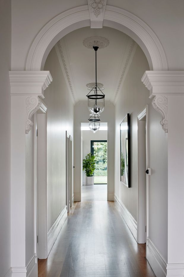 Elegant long residential hallway with polished timber flooring, white paneled walls, elaborate classical architraves, decorative corbels, ceiling mouldings, and suspended glass lantern lights. Multiple doorways and framed artwork line the corridor, which leads to a bright glazed opening and garden beyond. The space has a refined heritage character with a predominantly white and warm neutral palette.