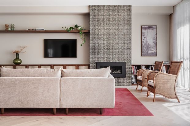 A bright, refined living room viewed from behind a large upholstered sectional sofa. The room features a full-height textured stone or mosaic fireplace surround with a dark built-in fireplace, integrated timber display shelving, a wall-mounted television, woven and timber lounge chairs, a red area rug, pale timber flooring, and sheer floor-to-ceiling curtains with strong natural light.