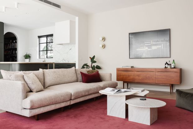 A refined open-plan living room with a large cream upholstered sectional sofa, sculptural white marble or stone coffee tables, a warm timber mid-century-style sideboard, and a deep red carpet. The space opens directly to a contemporary kitchen with dark cabinetry, a marble-look island or peninsula, black-framed glazing, and an arched dark shelving unit. Neutral walls, minimalist pendant-like wall lights, indoor plants, and framed artwork complete the polished interior.