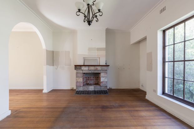 Bright, mostly empty traditional living room with polished dark timber flooring, white walls and decorative ceiling cornices. A brick and stone fireplace forms the focal point, while a large black-framed multi-pane window and arched opening add architectural character. An ornate chandelier hangs centrally from the ceiling.
