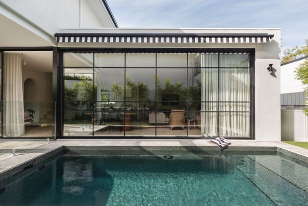 Contemporary white rendered home facade with a large black-framed glazed pavilion opening directly onto a tiled swimming pool. The glazing reveals a refined interior seating and dining area, with sheer curtains and garden reflections. A black-and-white striped awning adds a distinctive accent above the glass wall, while a narrow paved pool surround and adjacent white fencing complete the scene.