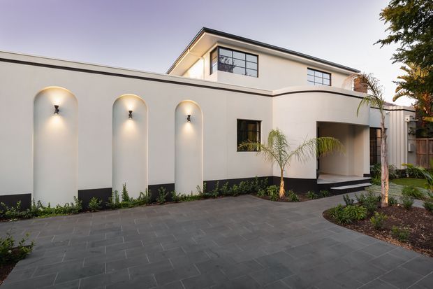 A contemporary two-storey residential exterior with smooth white rendered walls, strong black base detailing, recessed arched wall niches with sconces, dark-framed windows, and a covered entry. A broad dark grey paved forecourt, low planting, and slender palms frame the facade.