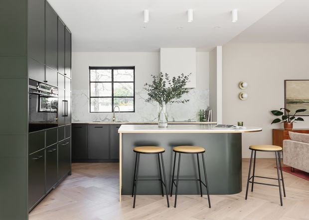 A contemporary open-plan kitchen with dark green-black floor-to-ceiling cabinetry, integrated appliances, a pale stone benchtop and matching curved island. Three timber-seat bar stools sit at the island, while a large black-framed window, pale herringbone timber flooring, indoor greenery and part of an adjacent living area are visible.