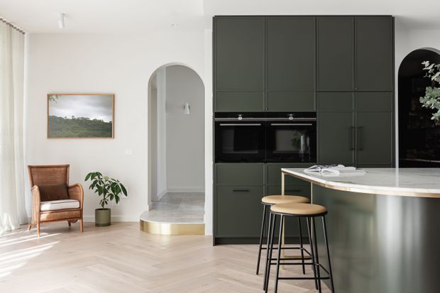 Bright open-plan kitchen and living area with a dark green, full-height built-in kitchen wall, integrated double ovens, curved light stone island benchtop, and two timber-and-black stools. The adjoining living area has pale timber flooring, white walls, sheer curtains, an upholstered armchair, potted plant, framed artwork, and an arched opening leading to a hallway. The space has a refined, contemporary feel with strong contrast between the dark cabinetry and light finishes.