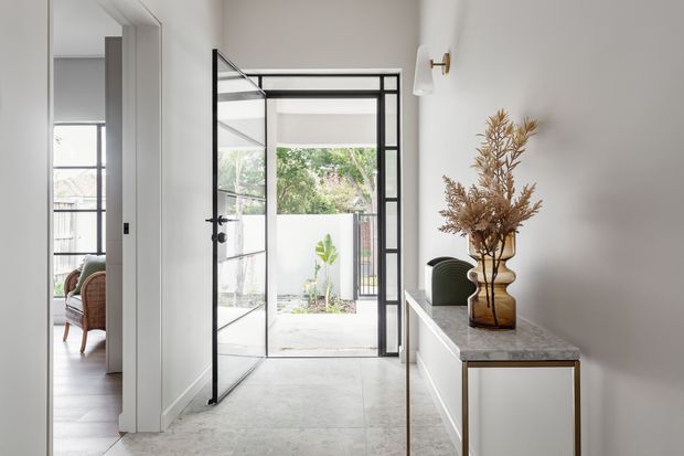 Bright contemporary entry hall with a pale stone or concrete-look floor, white walls, and a black-framed glazed steel door opening to a small courtyard. A slim white console with a stone top, decorative vase, wall sconce, and green upholstered chair define the space. A glimpse of a furnished room is visible through the doorway on the left.