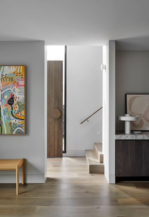 A refined contemporary interior focused on a compact entryway and stair hall. The space features pale grey walls, light timber flooring, a tall timber pivot door, minimalist stair detailing, and dark built-in cabinetry with a stone benchtop. Abstract artwork, a woven timber bench, and sculptural decor add warmth and colour. A stairway is visible beyond the entry, with a handrail mounted on the wall.