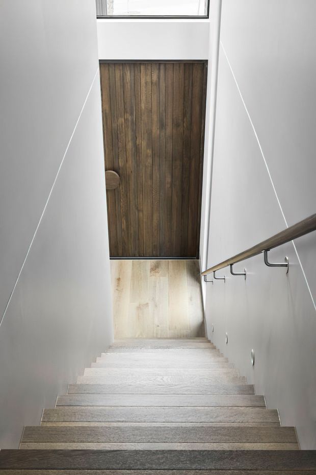 A narrow contemporary stairway viewed from above, with pale timber stair treads, white walls, a dark timber-clad door or feature panel at the lower landing, and a simple timber handrail with dark metal brackets. A high-level window brings in natural light.