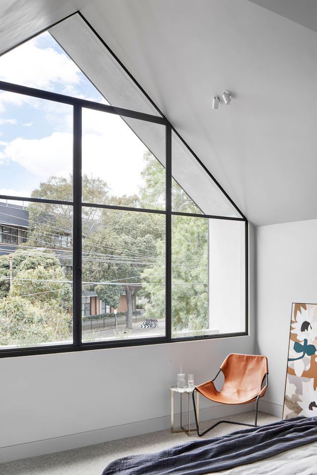Bright contemporary bedroom with a large black-framed, geometric floor-to-ceiling corner window beneath a sloping ceiling. The room has pale walls, light carpet, a low bed with dark bedding, and a sculptural tan leather lounge chair beside a small side table. Trees and neighbouring buildings are visible outside.