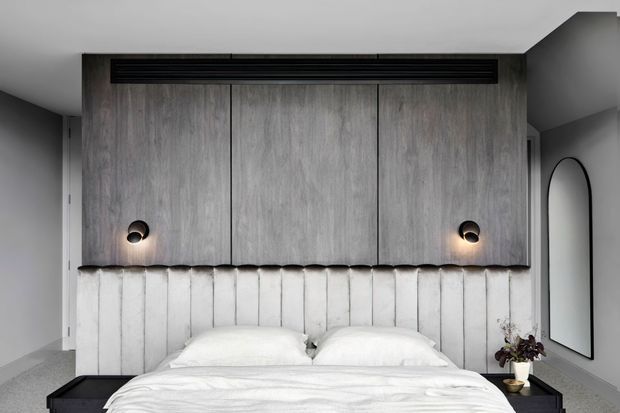 A contemporary bedroom with a low bed centered beneath a full-width built-in timber headboard and overhead cabinetry. The cabinetry has a weathered grey wood appearance, integrated dark linear ventilation or lighting detail, and two small wall lights. The lower headboard is upholstered in vertically channelled light fabric. The room has pale walls, dark grey ceiling and side accents, a large arched mirror, and minimal bedside styling.