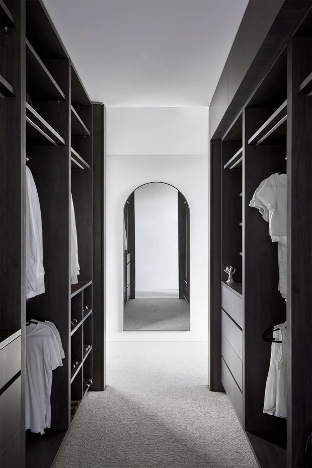 A narrow, refined walk-in wardrobe lined on both sides with dark timber built-in shelving, hanging rails, drawers and neatly arranged clothing. A full-height arched mirror is positioned at the end of the passage, with soft white walls and light grey carpet creating contrast against the dark joinery.