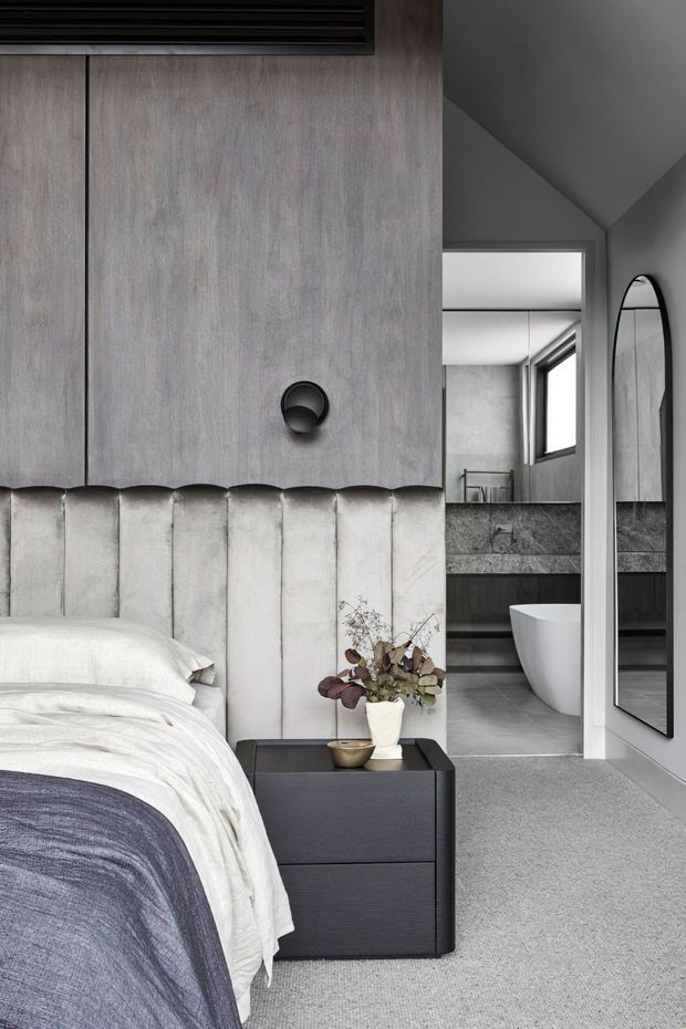 A refined contemporary bedroom with a partially visible bed, vertically channelled upholstered headboard, dark built-in cabinetry, and a compact black bedside table. A large rounded mirror reflects the adjacent ensuite bathroom, which contains a freestanding white bathtub, grey stone vanity, and dark cabinetry. The palette is predominantly soft grey, white, charcoal, and muted timber tones, with a pitched ceiling detail and plush carpet.