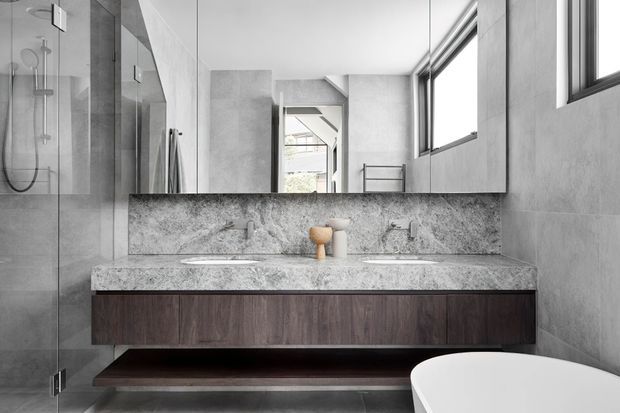 Contemporary bathroom with a floating dark timber double vanity, pale grey veined stone benchtop and integrated splashback, large wall mirror, frameless glass shower, and freestanding white bathtub. Grey wall and floor finishes create a refined monochromatic palette, with strong natural light from high windows.