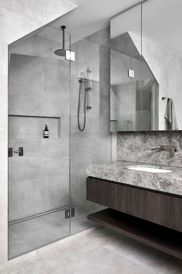 A contemporary bathroom featuring a frameless glass walk-in shower with rain showerhead and handheld fixture, large grey stone-look wall and floor tiles, a mirrored wall cabinet, and a floating dark timber vanity with a grey stone benchtop and integrated white basin. Brushed metal tapware and minimal detailing create a refined monochromatic scheme.