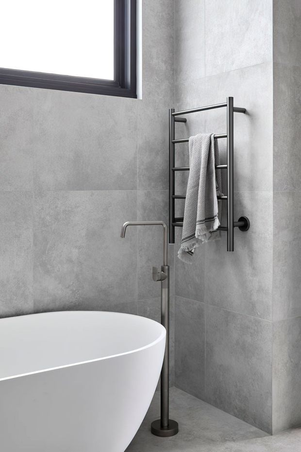 A contemporary bathroom corner featuring a freestanding white bathtub, a slim dark metal floor-mounted bath tap, large grey stone-look wall tiles, a matching grey tiled floor, and a dark metal heated towel rail with a draped towel. A black-framed window provides natural light.