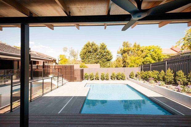 A contemporary covered outdoor entertaining area overlooking a rectangular swimming pool. The space features a timber-lined pergola roof with exposed beams, dark structural posts and framing, a large ceiling fan, dark tiled or stone paving, glass balustrades, and landscaped raised garden beds enclosed by dark fencing. Adjacent residential roofs and trees are visible beyond the pool.