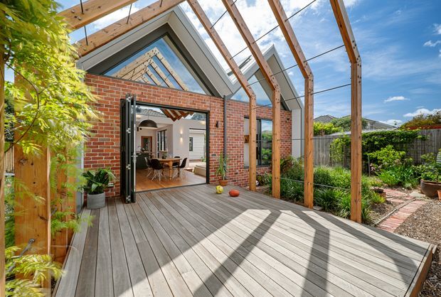 A bright residential outdoor deck or terrace beneath a timber-framed glazed pergola, attached to a brick extension with large bi-fold doors. The deck overlooks a planted garden and leads into a dining area visible through the open doors. Exposed timber framing, red brick walls, grey-toned decking, extensive glazing, and strong sunlight create an airy indoor-outdoor living space.