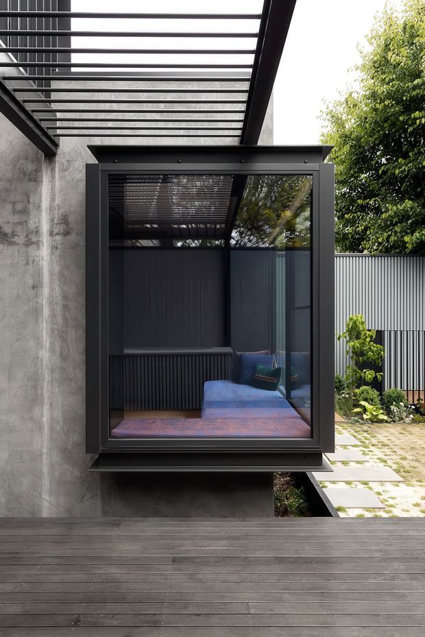 A contemporary residential exterior showing a dark timber deck, exposed concrete wall, black-framed projecting glass window or enclosed nook, and a covered pergola with translucent roofing. Through the glazing is a compact dark-clad seating area with a blue sofa and cushions. A small landscaped courtyard with patterned paving and corrugated metal fencing is visible to the right.
