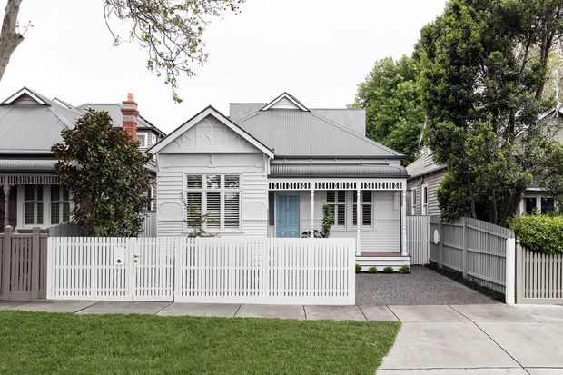 Street-facing view of a compact heritage-style single-storey residence with a white painted weatherboard facade, decorative gabled front section, grey corrugated metal roof, white picket fencing, plantation shutters, and a blue front door. A paved driveway and landscaped front verge are visible.
