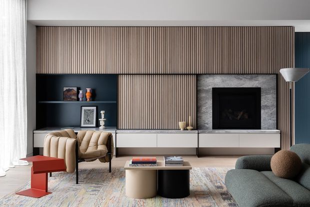 Contemporary living room featuring a full-width built-in media wall with vertical timber slat detailing, dark open shelving, a central concealed television panel, and a fireplace set within a grey stone-look surround. A large sectional sofa, sculptural coffee tables, lounge chairs, floor lamp, rug, and sheer curtains complete the refined interior.