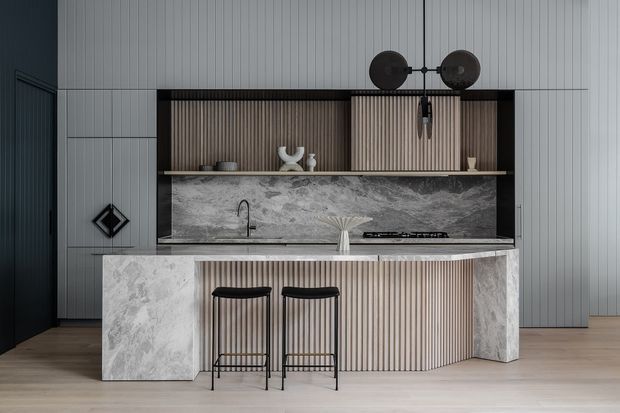 A sophisticated contemporary kitchen featuring a large marble-look island with waterfall ends and vertically fluted cabinetry. The island includes two black metal bar stools, while a matching textured backsplash and open shelving sit behind the cooktop and sink. Grey wall panelling, dark accents, pale timber flooring, and sculptural pendant lighting create a refined, monochromatic interior.