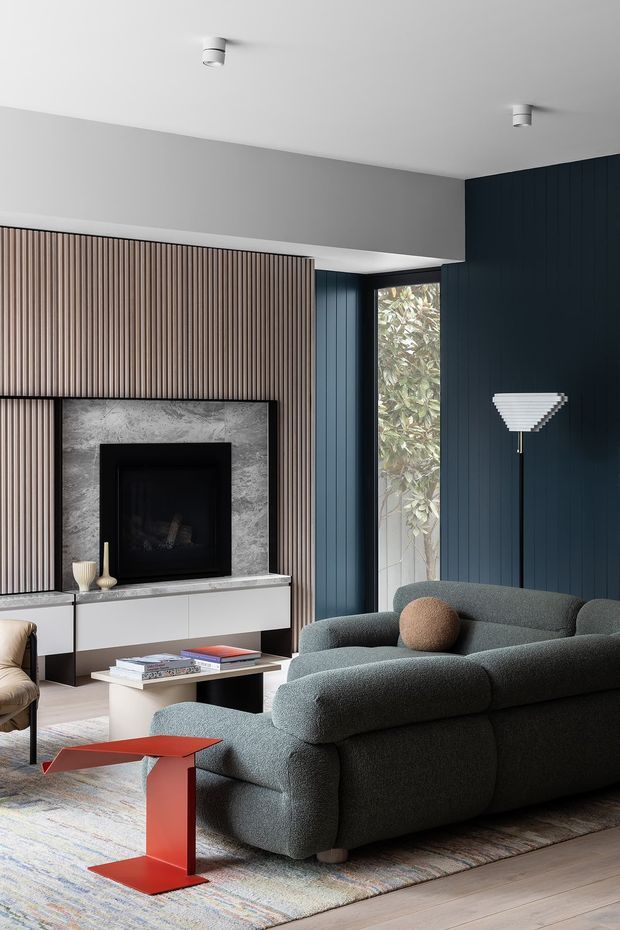 A refined contemporary living room centred on a fireplace, with vertically fluted timber wall panelling, dark blue vertical wall panelling, a grey upholstered sectional sofa, and a slim vertical window. The fireplace is framed in grey stone and integrated into white built-in cabinetry. A pale patterned rug, white coffee table, red side table, and sculptural wall lamp add contrast to the restrained interior.