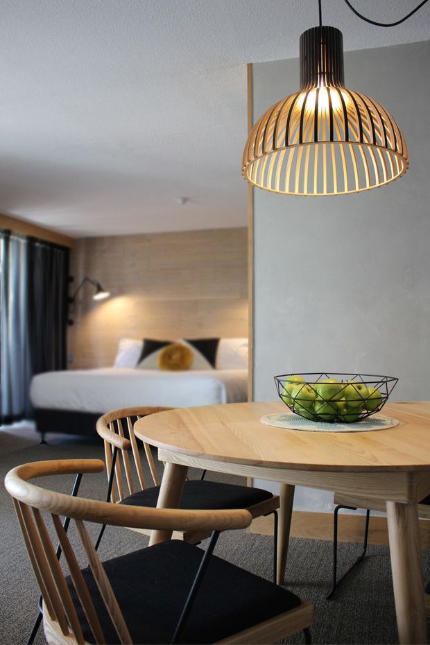 A warm, compact open-plan room combining a dining area and bedroom. In the foreground is a round light timber dining table with black upholstered timber-framed chairs, a wire fruit bowl, and a sculptural pendant light. Behind a partial dividing wall, a neatly made bed sits against a light timber feature wall, with dark curtains and a wall-mounted reading light. The space has a calm, Nordic-inspired palette of light timber, white, charcoal, and warm pendant lighting.