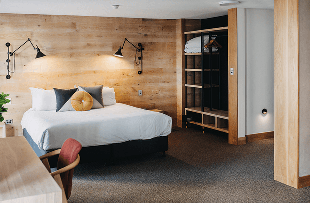 Warm, contemporary bedroom with a low-profile upholstered bed dressed in white linens and layered cushions, set against a full-height light timber feature wall. Black articulated wall sconces, open timber-and-black wardrobe shelving, a grey painted wall, and charcoal carpet create a refined, earthy interior. A timber desk or console and upholstered chair are partially visible in the foreground.