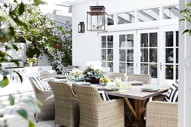Bright, elegant outdoor dining terrace with a large rustic timber dining table surrounded by woven rattan dining chairs. The space features white architectural detailing, extensive French-style glazed doors and windows, decorative lantern lighting, potted plants, and a built-in outdoor cooking area, creating a refined garden entertaining setting.