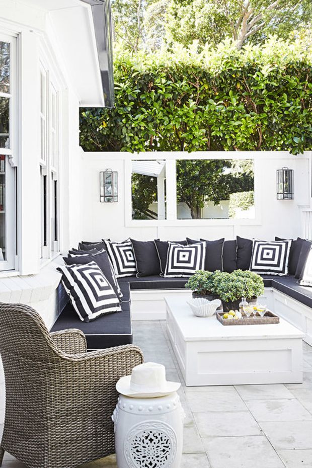 Bright white outdoor courtyard lounge featuring an L-shaped built-in bench with dark cushions and bold black-and-white geometric pillows, a white rectangular coffee table, woven wicker armchair, and decorative white ceramic stool. White rendered walls, framed openings, outdoor lanterns, pale stone paving, and dense green hedge planting create a crisp, sheltered entertaining space.