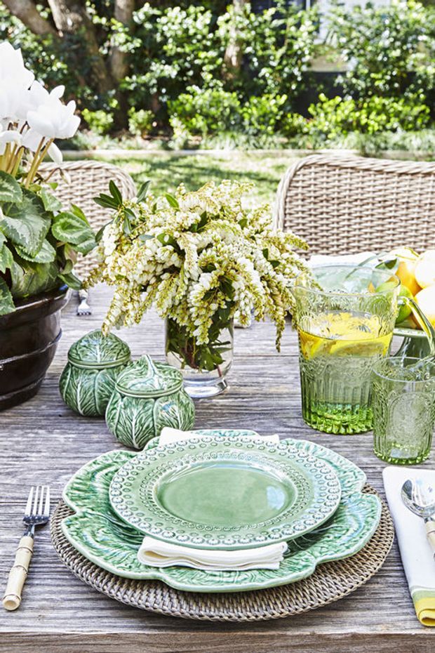Bright outdoor dining table setting in a leafy garden, featuring a weathered timber tabletop, woven wicker chairs and placemats, green patterned ceramic dinnerware, clear green-tinted glassware, citrus-infused water, and floral arrangements. The scene has a relaxed, elegant garden entertaining atmosphere.