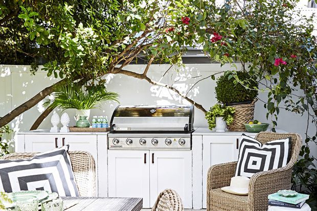 A bright outdoor entertaining area with a built-in stainless-steel barbecue set into white cabinetry, framed by lush foliage and flowering branches. Wicker armchairs with bold black-and-white geometric cushions surround a light timber outdoor table, creating a relaxed garden dining and cooking space.