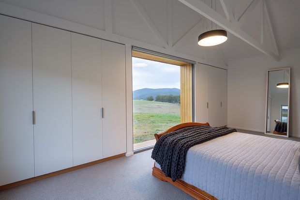 A bright, spacious bedroom with built-in white wardrobes, a timber-framed bed, soft grey bedding, carpeted flooring, and exposed white structural roof trusses. Full-height glazing or an open glass door provides views across a rural landscape and hills. A simple pendant light and full-length mirror complete the calm, minimalist interior.