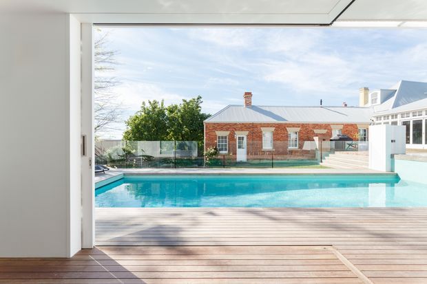 View from a modern interior through wide glazing toward a rectangular swimming pool, timber deck and a brick residential building. The scene features clean white interior surfaces, expansive glazing, a glass pool fence, and a bright contemporary outdoor setting.