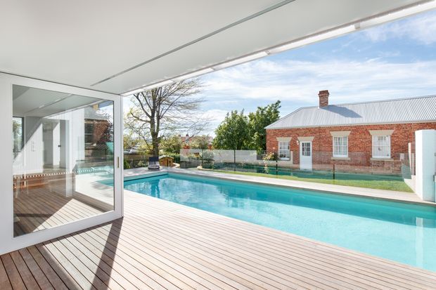 Bright architectural view from a glazed indoor/outdoor living space onto a large rectangular swimming pool and timber deck. A low red-brick, single-storey building with a corrugated metal gable roof is visible beyond the pool, along with established trees and a clear sky. The image emphasises indoor-outdoor connection, expansive glazing, and a clean contemporary poolside setting.