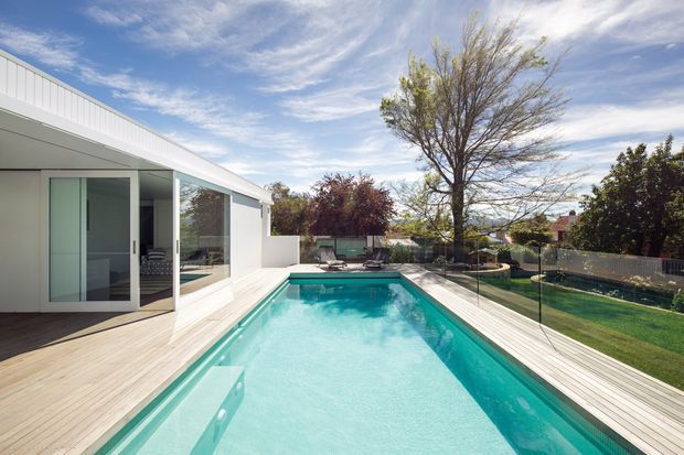 Bright contemporary residential pool area viewed from a timber deck beside a white-clad pavilion or extension. A long rectangular turquoise swimming pool is bordered by clear glass safety fencing and pale timber decking, with lawn, mature trees and neighbouring homes beyond. The building has clean angular rooflines, large glazing and minimal white cladding under a blue sky.