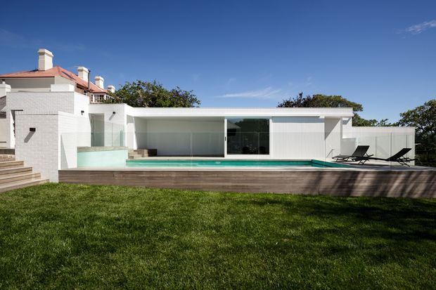 A low, modern white-rendered pool pavilion with extensive floor-to-ceiling glazing, a long turquoise swimming pool, and a timber-clad raised deck. The pavilion connects to a larger light-coloured residence at the left, with outdoor loungers beside the pool and a broad lawn in the foreground.
