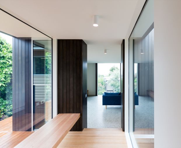 A bright contemporary interior passage or entry zone with extensive floor-to-ceiling glazing, dark vertical timber wall panelling, and a light timber bench or console. The view looks through to a living room with a blue sofa and carpeted floor, while lush greenery is visible outside through the glass. Clean-lined recessed and surface lighting, pale walls, and reflective glazing create an airy, modern connection between interior and garden.