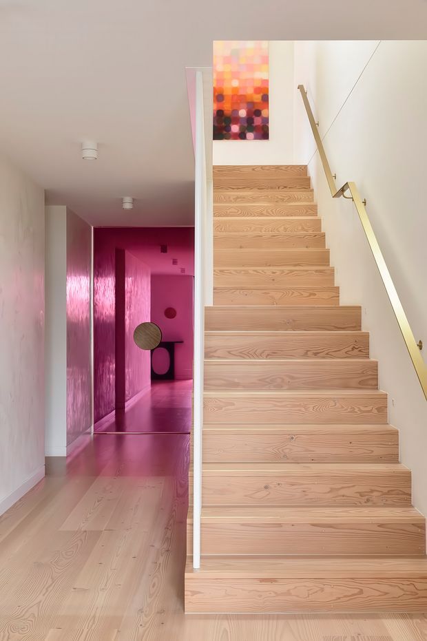 Bright contemporary interior hallway and stairway with a broad light timber staircase, pale timber flooring, white walls, and a slim brass-toned handrail. A vivid pink/magenta corridor extends to the left, creating a strong colour contrast, while a colourful abstract artwork is mounted above the stairs.