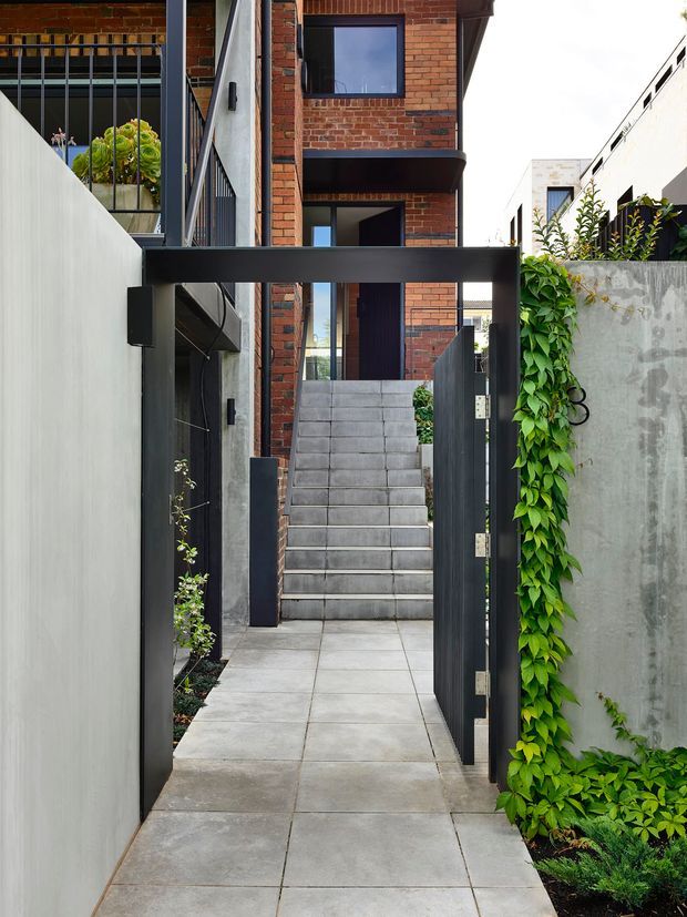 A narrow contemporary residential entry passage with large square concrete pavers leading through a dark metal gate toward a raised staircase. The passage is enclosed by smooth pale rendered walls, exposed red-brick apartment-style facades, black metal screening and a raw concrete boundary wall softened by climbing greenery. The composition emphasises the layered threshold, stairs and vertically compact urban architecture.