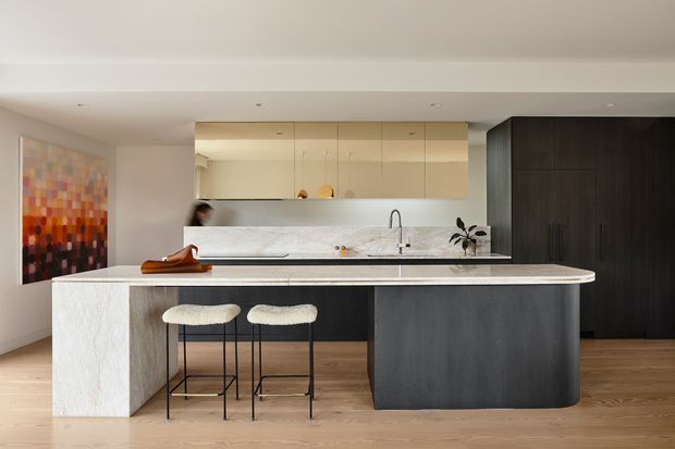 Contemporary open-plan kitchen with a large marble-clad island, dark timber cabinetry, pale wood flooring, and a minimalist seating area. Light upper cabinetry and a marble splashback create a clean, refined interior, with colourful wall art adding a subtle accent.