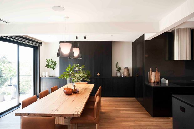 Contemporary open-plan dining and kitchen interior with a long solid timber dining table, brown upholstered dining chairs, dark built-in cabinetry, black benchtops, pendant lighting, timber flooring, and full-height glazed doors opening to an outdoor area. Indoor plants and warm timber tones soften the high-contrast black-and-white scheme.