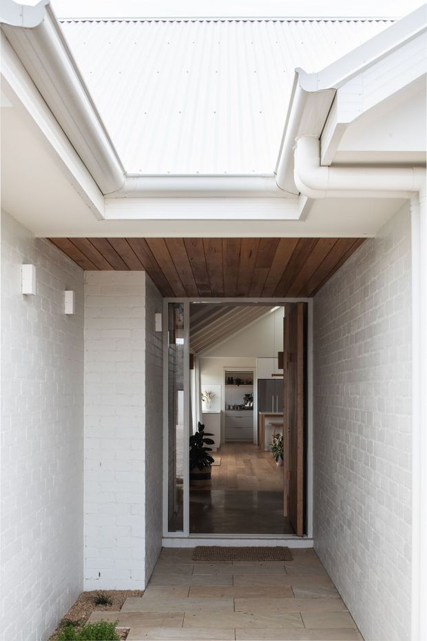 A bright covered entry passage leading to a glazed timber-framed front door. White-painted brick walls and white metal roofing with exposed gutters surround a warm timber-lined ceiling. Through the doorway, a calm interior with timber flooring, a pitched ceiling, kitchen joinery and indoor plants is visible.