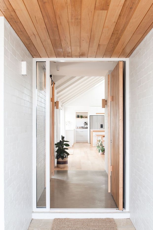 Bright architectural photograph looking through a narrow entry passage into an open-plan kitchen and living area. The entry has white-painted brick walls, a warm timber-lined ceiling, a polished concrete floor, and a large timber-framed glazed door with timber panels. Beyond the doorway is a light-filled interior with pale timber flooring, white cabinetry, exposed sloping roof beams, and potted plants.