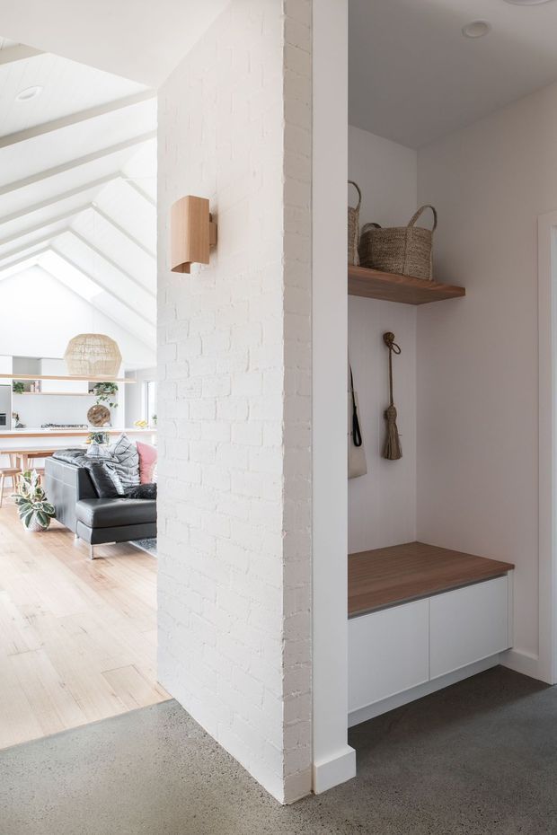 Bright residential entryway with a built-in mudroom nook, featuring white-painted brick walls, a timber bench and shelf, white lower cabinetry, hooks, woven baskets, and carpeted flooring. The entry opens toward an airy living room with pale timber floors, a dark sectional sofa, and a glazed or skylit roof structure. A compact timber wall sconce is mounted on the brick pier.