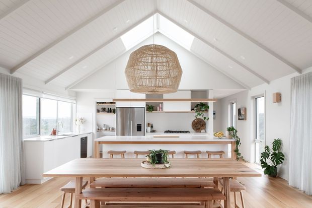 Bright open-plan kitchen and dining area with a white vaulted timber-lined ceiling, exposed beams, clerestory glazing, and light timber flooring. A large light-wood dining table and bench seating sit in the foreground, while the rear kitchen features a white island, timber accents, open shelving, stainless-steel appliances, and a woven pendant light. Full-height windows and sheer curtains create an airy, relaxed interior.