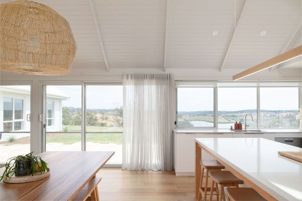 Bright open-plan kitchen and dining area with extensive floor-to-ceiling glazing overlooking a green rural landscape and water. The room features pale timber flooring, white cabinetry, a large white island with timber trim and stools, a timber dining table, sheer curtains, and a woven pendant light. White painted timber-lined walls and a pitched ceiling create an airy coastal-residential feel.