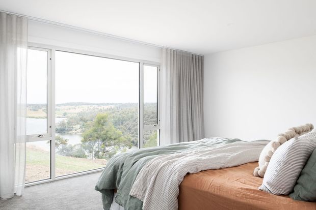 Bright, airy bedroom with floor-to-ceiling glazing overlooking a leafy landscape and water. The room features a low-profile bed with layered sage green, white, and soft brown bedding, a plush textured headboard, sheer white curtains, grey drapes, light carpet, and clean white walls.